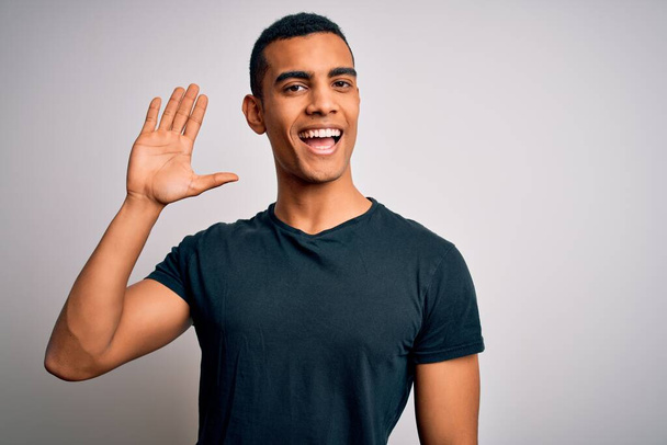 Joven hombre afroamericano guapo con camiseta casual de pie sobre fondo blanco Renuncia a decir hola feliz y sonriente, gesto de bienvenida amistoso
 - Foto, imagen
