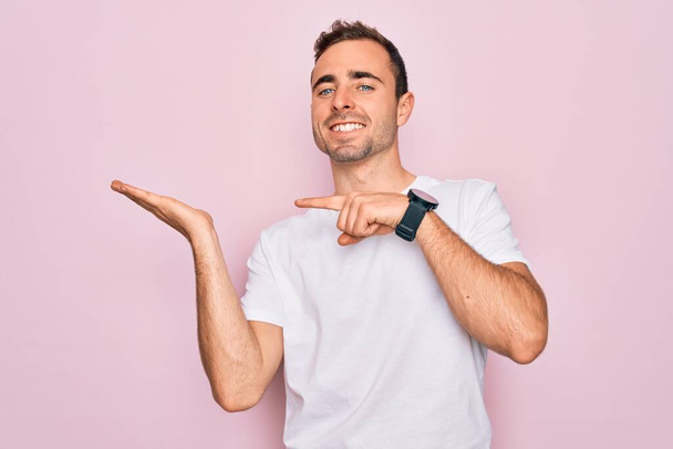 Handsome man with blue eyes wearing casual white t-shirt standing over pink background amazed and smiling to the camera while presenting with hand and pointing with finger. - Photo, Image
