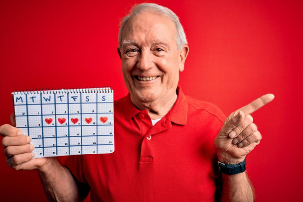 Senior hombre de pelo gris con calendario de fecha especial sobre fondo rojo muy feliz señalando con la mano y el dedo a un lado
 - Foto, imagen