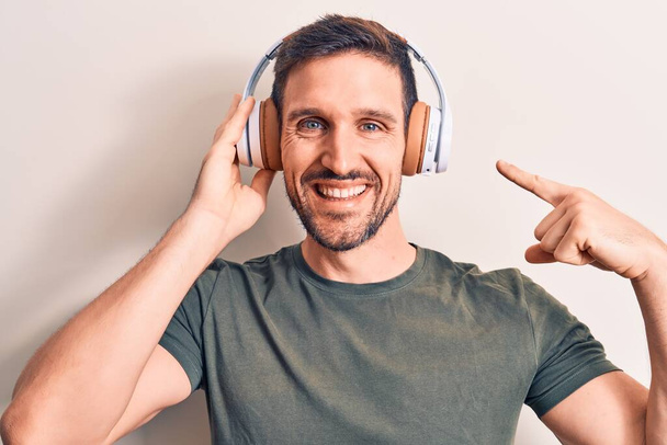 Nuori komea mies kuuntelee musiikkia käyttämällä kuulokkeita yli eristetty valkoinen tausta hymyillen onnellinen osoittaa käsin ja sormella - Valokuva, kuva