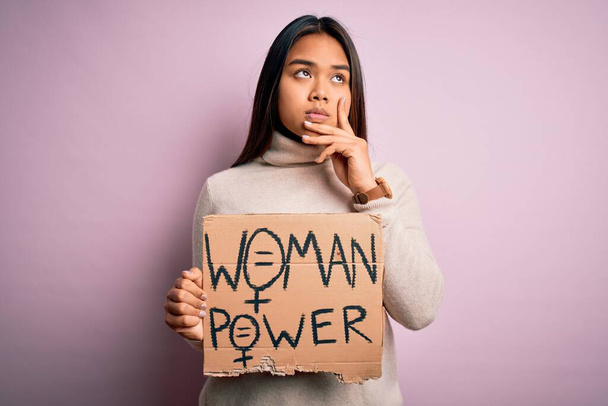 Giovane ragazza attivista asiatica chiede diritti delle donne tenendo banner con la donna messaggio di potere volto serio pensando alla domanda, idea molto confusa - Foto, immagini
