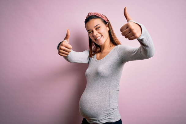 Jong mooi tiener meisje zwanger verwacht baby over geïsoleerde roze achtergrond goed te keuren doen positieve gebaar met de hand, duimen omhoog glimlachend en blij voor succes. Winnaar gebaar. - Foto, afbeelding