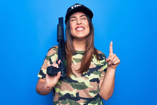 Young beautiful hunter woman wearing camouflage t-shirt and usa cap holding shotgun smiling with an idea or question pointing finger with happy face, number one - Фото, зображення