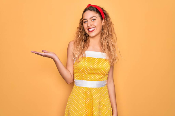 Beautiful blonde pin-up woman with blue eyes wearing diadem standing over yellow background smiling cheerful presenting and pointing with palm of hand looking at the camera. - Fotó, kép