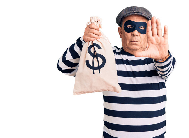 Senior όμορφος άντρας φορώντας μάσκα διαρρήκτη κρατώντας τσάντα χρήματα με ανοιχτό χέρι κάνει στοπ υπογράψει με σοβαρή και σίγουρη έκφραση, χειρονομία άμυνας  - Φωτογραφία, εικόνα