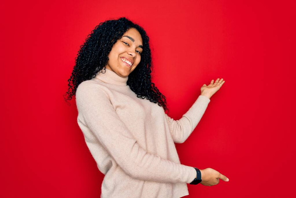 Joven mujer rizada afroamericana con suéter casual cuello alto sobre fondo rojo invitando a entrar sonriente natural con la mano abierta
 - Foto, imagen