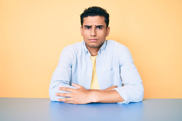 Nuori komea latinomies päällään rento vaatteita istuu pöydällä skeptinen ja hermostunut, paheksuva ilme kasvot ristissä kädet. negatiivinen henkilö.  - Valokuva, kuva