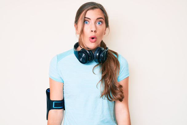 Beautiful young caucasian woman wearing gym clothes and using headphones afraid and shocked with surprise expression, fear and excited face.  - Photo, Image