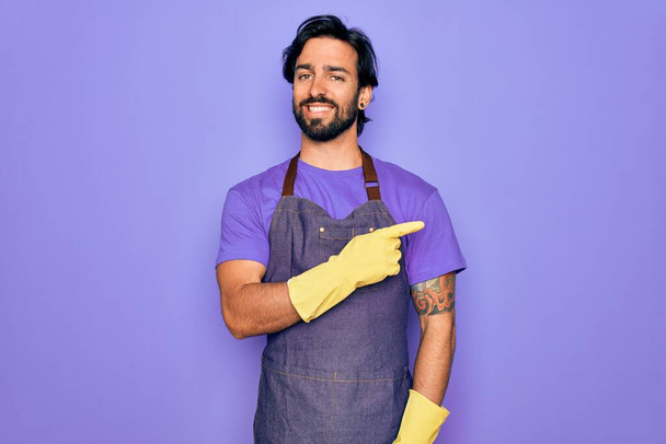 Fiatal jóképű spanyol clenaer férfi visel házimunka kötény és mosás kesztyű vidám egy mosoly az arc mutat kézzel és ujjal oldalra boldog és természetes kifejezés az arcon - Fotó, kép