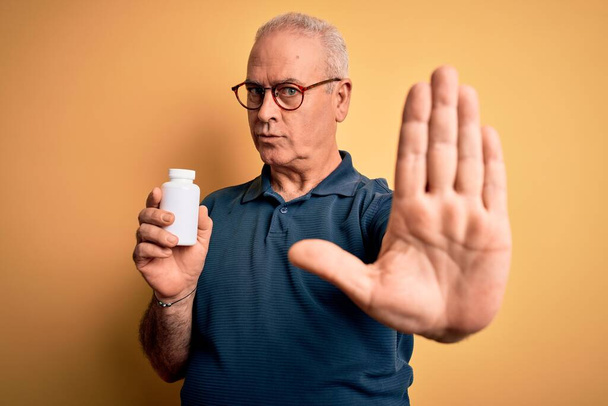 Чоловік середнього віку тримає ліки таблетки стоячи над ізольованим жовтим тлом з відкритою рукою робить знак зупинки з серйозним і впевненим виразом, захисний жест
 - Фото, зображення