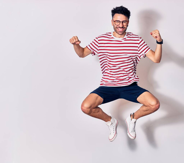 Young handsome man wearing casual clothes and glasses smiling happy. Jumping with smile on face doing winner sign with fists up over isolated white background - Fotó, kép