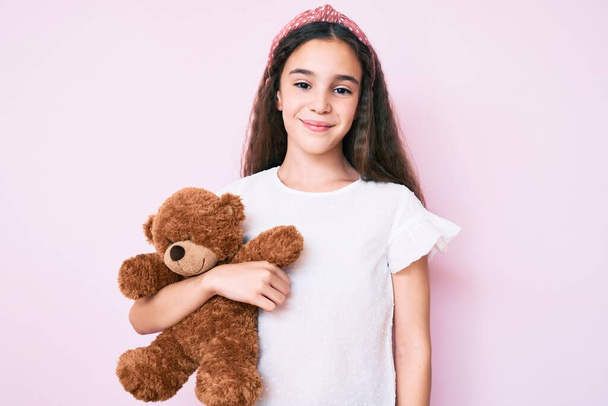 Schattig hispanic kind meisje houden teddy beer kijken positief en gelukkig staan en glimlachen met een zelfverzekerde glimlach tonen tanden  - Foto, afbeelding