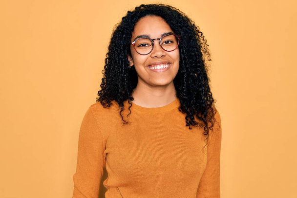 Nuori afrikkalainen amerikkalainen nainen yllään rento villapaita ja lasit keltaisella taustalla onnellinen ja viileä hymy kasvoilla. Onnekas henkilö. - Valokuva, kuva