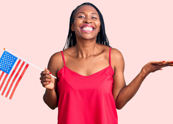 Νεαρή Αφροαμερικανή γυναίκα που κρατά σημαία των Ηνωμένων Πολιτειών γιορτάζει τη νίκη με χαρούμενο χαμόγελο και έκφραση νικητή με υψωμένα χέρια  - Φωτογραφία, εικόνα