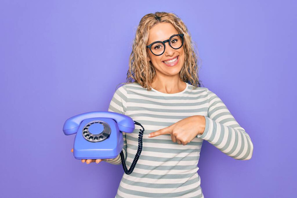 Jovem mulher loira bonita segurando telefone vintage sobre fundo roxo isolado sorrindo feliz apontando com a mão e o dedo - Foto, Imagem
