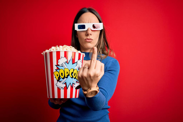 Joven mujer rubia con gafas 3D y comer paquete de palomitas de maíz viendo una película en el cine Mostrando el dedo medio, descortés y grosero joder la expresión
 - Foto, Imagen