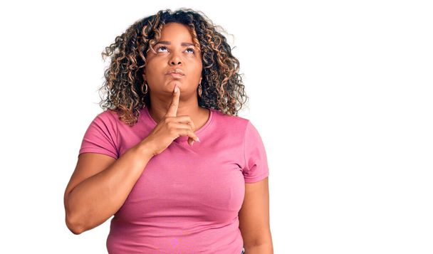 Junge afrikanisch-amerikanische Plus-Size-Frau in lässiger Kleidung, die konzentriert mit dem Finger am Kinn über Zweifel nachdenkt und fragend nach oben schaut  - Foto, Bild