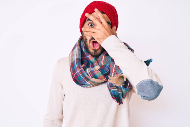 Красивый молодой человек с кудрявыми волосами и медведем в зимнем свитере, шарфе и шерстяной шляпе, заглядывающий рукой в лицо и глаза, испуганно смотрящий сквозь пальцы
  - Фото, изображение