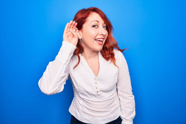 Jonge mooie roodharige zakenvrouw met een elegant shirt over een blauwe achtergrond die glimlacht met hand op hand luisterend en horend naar geruchten of roddels. Doofheid. - Foto, afbeelding