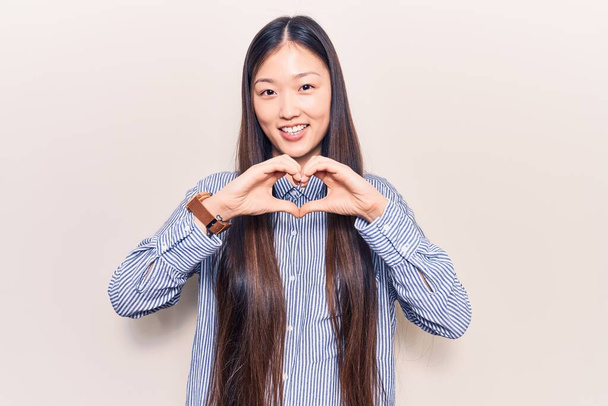 Genç, güzel Çinli bir kadın, sıradan çizgili tişört giyiyor, aşıkken gülümsüyor, elleriyle kalp sembolü yapıyor. romantik kavram.  - Fotoğraf, Görsel
