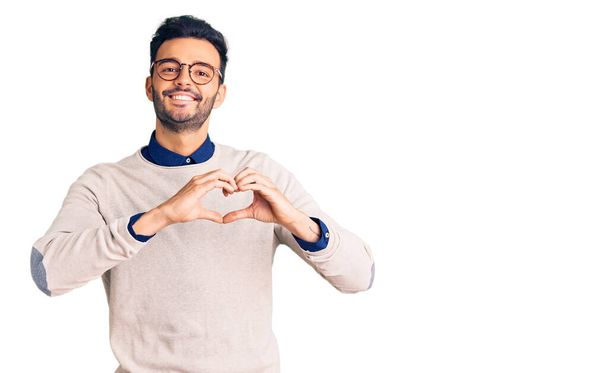 Jeune homme hispanique beau portant des vêtements élégants et des lunettes souriant en amour faisant forme de symbole de coeur avec les mains. concept romantique.  - Photo, image