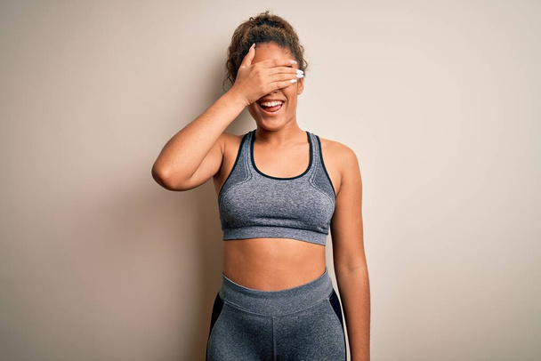 Jonge Afrikaanse amerikaanse sportvrouw die sport beoefent met sportkleding over een witte achtergrond glimlachend en lachend met de hand op het gezicht ogen bedekkend voor verrassing. Blind concept. - Foto, afbeelding