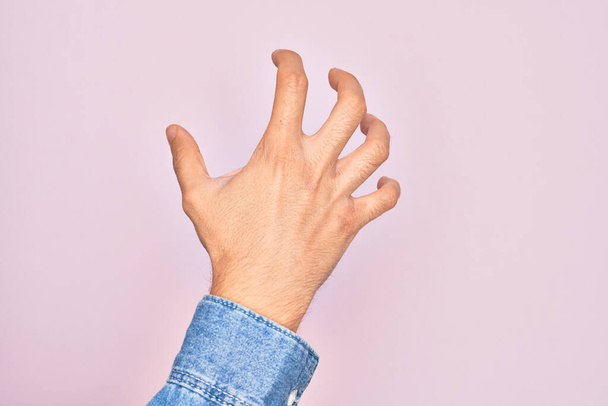 Hand van Kaukasische jonge man met vingers over geïsoleerde roze achtergrond grijpen agressief en eng met vingers, geweld en frustratie - Foto, afbeelding