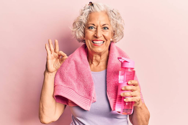 Старшая седовласая женщина в спортивной одежде и полотенце, пьющая бутылку воды делает хорошо знак с пальцами, улыбаясь дружелюбный жест отличный символ  - Фото, изображение