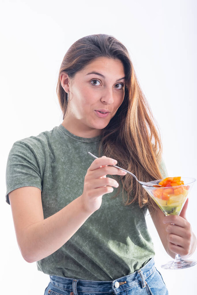 Mulher bonita segurando um copo de martini com pedaços de frutas sortidas e uma colher enquanto olha para a câmera em um fundo claro. Conceito de comida saudável e diversão
. - Foto, Imagem