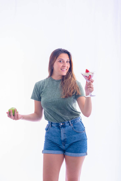Beautiful young woman holding a martini glass with strawberries and whipped cream and a green apple while smiling at the camera on a light background. Healthy desserts and fun concept. - Фото, изображение