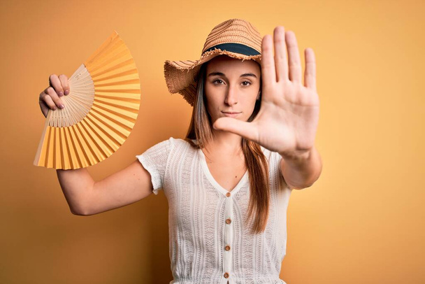 Yaz şapkası takan genç turist kadın sarı arka planda el ele tutuşmuş yelpaze açık elle dur işareti yapıyor ciddi ve kendinden emin bir ifadeyle, savunma jesti yapıyor. - Fotoğraf, Görsel