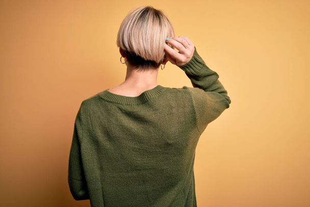 Young blonde woman with modern short hair wearing casual sweater over yellow background Backwards thinking about doubt with hand on head - Foto, imagen