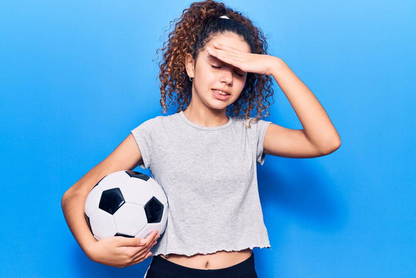Красивая девочка с вьющимися волосами держа футбольный мяч напряжены и разочарованы с рукой на голове, удивлен и сердитый лицо  - Фото, изображение