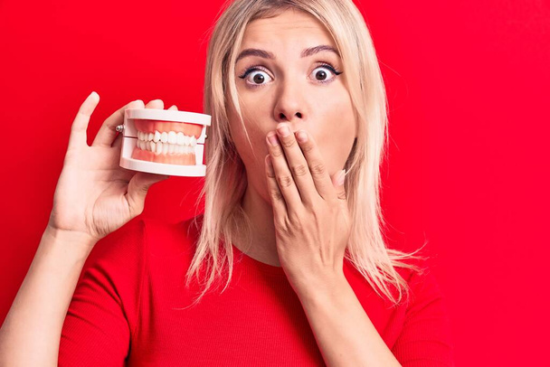 Young beautiful blonde woman holding denture teeth standing over isolated red background covering mouth with hand, shocked and afraid for mistake. Surprised expression - Photo, Image