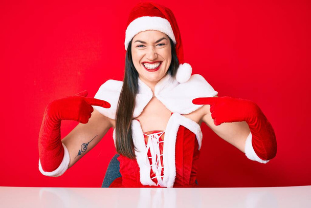 Молодая белая женщина в костюме Санта-Клауса выглядит уверенно с улыбкой на лице, указывая на себя пальцами гордой и счастливой.  - Фото, изображение