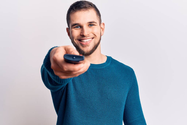 Young handsome man holding tv remote control looking positive and happy standing and smiling with a confident smile showing teeth  - Photo, image