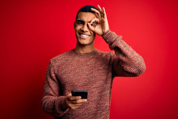 Yakışıklı Afro-Amerikalı adam kırmızı arka planda akıllı telefon kullanarak konuşuyor. Gülümsüyor. Elleri gözlerinde, parmaklarının arasından bakıyor. - Fotoğraf, Görsel