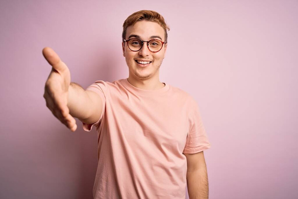 Joven hombre pelirrojo guapo con camiseta casual de pie sobre fondo rosa aislado sonriendo amistoso ofreciendo apretón de manos como saludo y bienvenida. Negocios exitosos
. - Foto, imagen