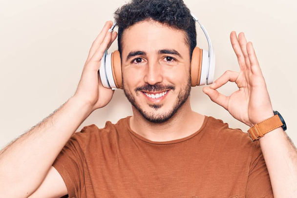 Jeune homme hispanique écoutant de la musique à l'aide d'écouteurs faire ok signe avec les doigts, sourire geste amical excellent symbole  - Photo, image