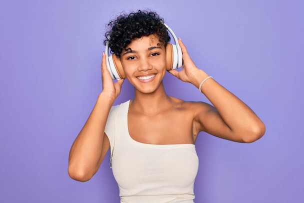 Mulher americana africana bonita ouvindo música usando fones de ouvido sobre fundo vermelho com um rosto feliz de pé e sorrindo com um sorriso confiante mostrando dentes
 - Foto, Imagem