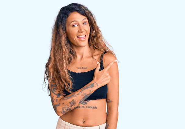 Mujer hispana joven con tatuaje usando ropa casual alegre con una sonrisa de la cara señalando con la mano y el dedo hacia arriba al lado con expresión feliz y natural en la cara
  - Foto, imagen