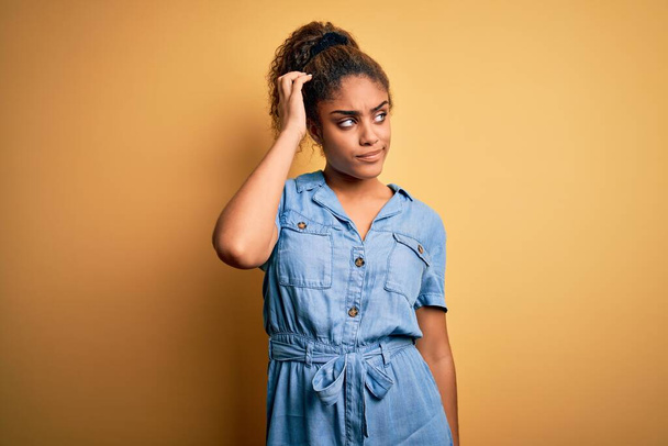 Mladý krásný africký americký dívka na sobě džínové šaty stojící nad žlutým pozadím matoucí a přemýšlel o otázce. Nejistý pochybami, přemýšlející s rukou na hlavě. Pensive concept. - Fotografie, Obrázek