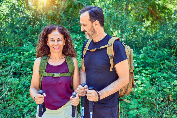 Beautiful couple of hiker wearing backpack smiling happy. Standing with smile on face doing trekking using hiking stick at forest - Photo, Image