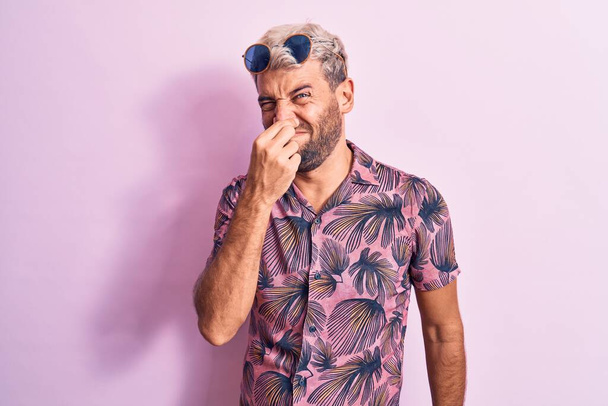 Schöner blonder Mann im Urlaub mit lässigem Hemd und Sonnenbrille über rosa Hintergrund, der etwas Stinkiges und ekelhaften, unerträglichen Geruch riecht und mit den Fingern auf der Nase den Atem anhält. Schlechter Geruch - Foto, Bild