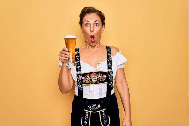 Középkorú barna nő visel német hagyományos oktoberfest ruha ivás pohár sört megijedt a sokk egy meglepetés arc, fél, és izgatott a félelem kifejezés - Fotó, kép