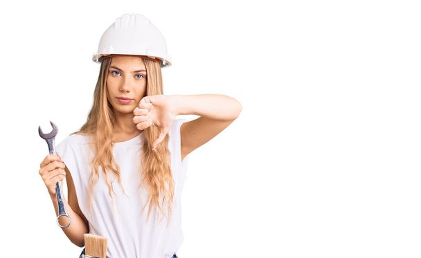 Beautiful caucasian woman with blonde hair wearing hardhat and builder clothes with angry face, negative sign showing dislike with thumbs down, rejection concept  - Photo, Image