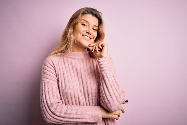 孤立した背景にカジュアルなピンクのセーターを着た若い美しいブロンドの女性は、交差した腕と手で笑顔でカメラに自信を持って見えます顎に提起.肯定的な考え. - 写真・画像