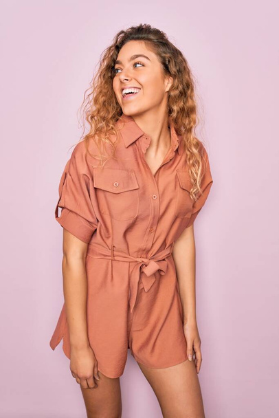 Young beautiful blonde woman with curly hair smiling happy and confident. Standing with smile on face wearing casual dress over isolated pink background - Fotografie, Obrázek