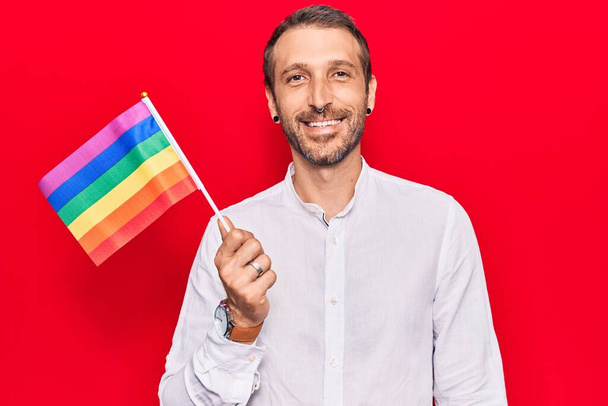 Joven hombre guapo sosteniendo arco iris lgbtq bandera buscando positivo y feliz de pie y sonriendo con una sonrisa confiada mostrando los dientes
  - Foto, imagen
