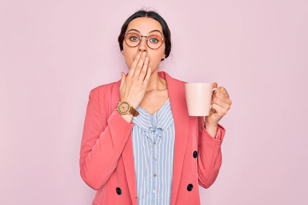 Young beautiful businesswoman with blue eyes wearing glasses drinking pink mug of coffee cover mouth with hand shocked with shame for mistake, expression of fear, scared in silence, secret concept - Foto, afbeelding